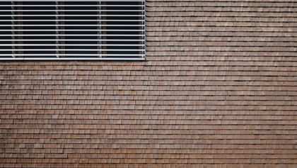 Holzfassade eines Gebäudes mit einem Fenster, an dem horizontale Lamellen angebracht sind.