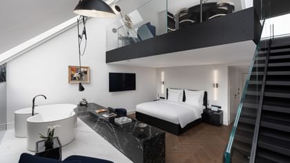 Zweistöckige Grand-Maisonette-Suite im „The Amauris“ Wien mit freistehender weißer Designerbadewanne auf Marmor, Doppelbett mit weißer Bettwäsche, wandmontiertem Flachbildfernseher und schwarzer Treppe mit Glasgeländer zu einer offenen Galerie mit Sitzecke.