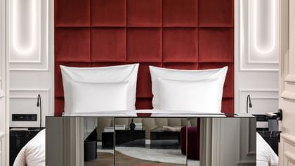 Elegantes Schlafzimmer der Opera Suite im „The Amauris“ Wien mit hohem bordeauxrotem Samtkopfteil hinter einem Doppelbett, großem Glaslüster, verspiegeltem Sideboard, rundem Samthocker und hellem Parkettboden.