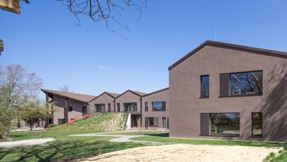 Zweigeschossiges Gebäude mit brauner Fassade und großen Fenstern, umgeben von einer grünen Wiese und Bäumen. Im Vordergrund ist ein Sandbereich sichtbar.
