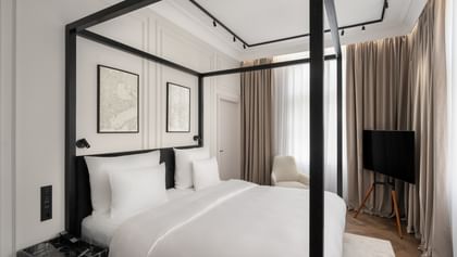 Helles Executive-Suite-Schlafzimmer im „The Amauris“ Wien mit modernem Himmelbett in schwarzem Rahmen, weißer Bettwäsche, abstrakten Wandbildern, bodentiefen Vorhängen, Lounge-Sessel und freistehendem TV auf hellem Parkett.