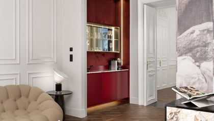 Wohnbereich der Opera Suite im „The Amauris“ Wien mit organisch geformtem beigen Sessel, gläsernen Couchtischen mit Bildbänden, Sideboard mit Steinfront, moderner Tischleuchte und tiefroter Nische mit beleuchteter Bar und Kaffeemaschine.