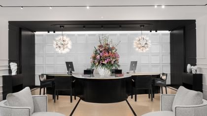 Helle Hotellobby des „The Amauris“ Wien mit halbkreisförmiger dunkler Rezeptionstheke, großem Blumenarrangement, drei Arbeitsplätzen vor kassettierten weißen Wänden, zwei kugelförmigen Glasleuchten und zwei gepolsterten Lounge-Sesseln im Vordergrund.