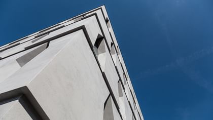 Die Ecke eines Gebäudes, die in die Höhe ragt. Die Fassade ist aus hellem Beton mit schmalen Fenstern, vor einem klaren blauen Himmel.