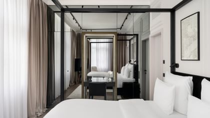 Helles Executive-Suite-Zimmer im „The Amauris“ Wien mit großem Doppelbett, weißer Bettwäsche, Spiegelfront mit Schreibtisch und schwarzem Stuhl, bodentiefen Vorhängen und gemütlicher Sitzecke mit Sessel und TV im Hintergrund.