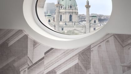 Loft-Suite im „The Amauris“ Wien mit rundem Bullaugenfenster und Blick auf die barocke Kuppel und Säulen der Karlskirche, davor eine grüne Samt-Eckcouch vor einer Wand mit großflächiger Architekturzeichnung.
