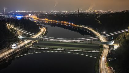 Eine nächtliche Luftaufnahme einer Brücke, die über einen Fluss führt. Die Brücke ist beleuchtet und reflektiert im Wasser.