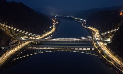 Eine nächtliche Luftaufnahme einer Brücke, die über einen Fluss führt. Die Brücke ist beleuchtet und reflektiert im Wasser.