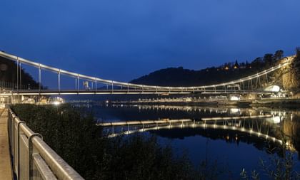 Eine nächtliche Luftaufnahme einer Brücke, die über einen Fluss führt. Die Brücke ist beleuchtet und reflektiert im Wasser.