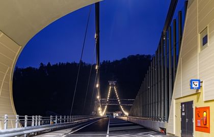 Eine nächtliche Aufnahme einer Brücke, mit gerader Sicht auf die Fahrbahn. Die Brücke ist beleuchtet, dahinter die Silhouette einer Hügelkette