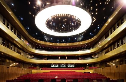  Innenansicht des Musiktheaters mit mehreren Ebenen von roten Sitzplätzen und einer auffälligen, runden Deckenleuchte
