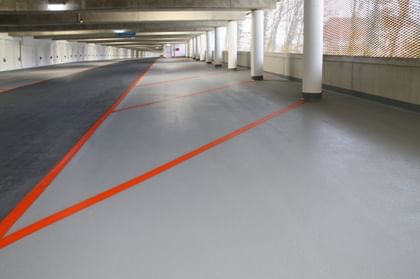 Der Boden einer Parkgarage ist in einem hellen Grauton gestrichen, während orangefarbene Linien diagonal über den Boden verlaufen, um Parkplätze oder Verkehrswege zu kennzeichnen. 
