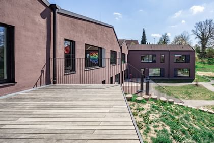 Braunes Gebäude mit einer flachen Holzterrasse im Vordergrund. Im Hintergrund sind grüne Flächen und Bäume zu sehen