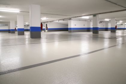Ein leerer Parkhausbereich mit hellen Wänden und einem grauen Boden. Die Wände sind teilweise mit blauen Streifen dekoriert. Es gibt mehrere Säulen und einige Feuerlöscher an den Wänden.