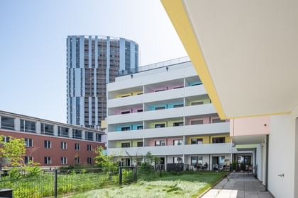 Mehrstöckiges Wohnhaus mit bunter Fassade, weißen Balkonen, Rasen und Bäumen, Hochhaus im Hintergrund