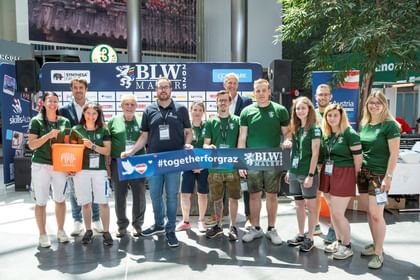 Eine Gruppe von Menschen steht an einem Veranstaltungsort, umgeben von Bannern und Sponsorenlogos. Sie tragen grüne T-Shirts und lächeln in die Kamera. In der Mitte hält eine Person ein Banner mit der Aufschrift „#togetherforgraz“