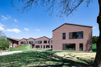 Zweigeschossiges Gebäude mit brauner Fassade und großen Fenstern, umgeben von einer grünen Wiese und Bäumen. Im Vordergrund ist ein Sandbereich sichtbar.