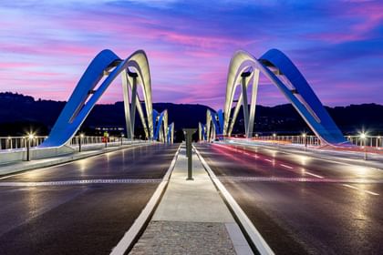Brücke mit geschwungenen, weißen Bögen, die sich über eine Straße erstreckt. Der Himmel ist in sanften Lila- und Rosatönen gefärbt.
