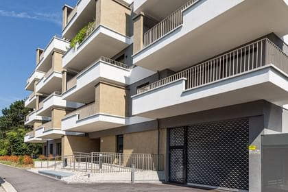 Wohnanlage in der Robert-Stolz-Straße in Linz. Ein modernes, mehrstöckiges Wohngebäude mit hellen Fassaden und zahlreichen Balkonen. Große Fenster sorgen für viel Tageslicht. Vor dem Gebäude stehen Autos auf einer ruhig wirkenden Straße, eingerahmt von gepflegten Grünflächen und Bäumen.