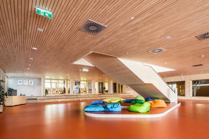 Ein Raum in einer Volkshochschule. Im Vordergrund sind bunte Sitzkissen in verschiedenen Farben (grün, blau) auf einem hellen Boden angeordnet. Auf der linken Seite ist eine Treppe, die zu einem oberen Bereich führt.
