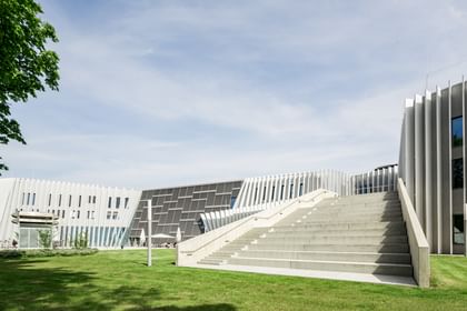 Modernes Gebäude der Anton Bruckner Privatuniversität mit markanter Fassadengestaltung aus vertikalen und horizontalen Lamellen. Eine breite Treppe führt zum Eingang, im Vordergrund befindet sich eine gepflegte Grünfläche.