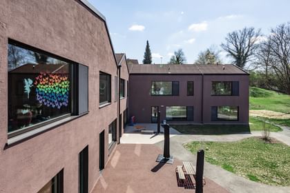 Zweigeschossiges Gebäude mit großen Fenstern und schwarzen Fensterläden. In den oberen Fenstern sind bunte Zeichnungen sichtbar.