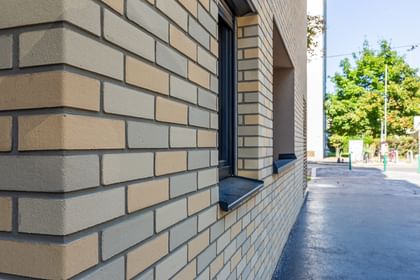 Nahaufnahme Fassade in Klinkeroptik mit Fenstern, davor asphaltierter Gehweg und Baum im Hintergrund