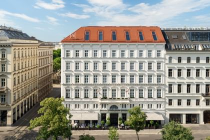 Außenansicht des „The Amauris“ Hotels in Wien mit klassizistischer weißer Fassade, symmetrischen Fenstern, rotem Mansarddach, Straßenbäumen im Vordergrund und blauem Himmel.