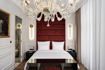 Schlafzimmer der Opera Suite im „The Amauris“ Wien mit großem Doppelbett vor hohem, weinrotem Kopfteil, vertikalen Lichtleisten in weißen Wandpanelen, opulentem Glaslüster und Einblick in ein marmorverkleidetes Badezimmer.