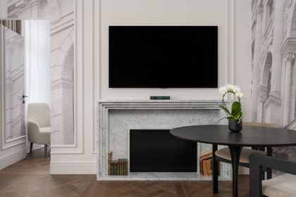 Wohnbereich einer Suite im „The Amauris“ Wien mit Kaminfront aus hellem Marmor, darüber ein Flachbildfernseher, runder Tisch mit zwei Stühlen und weißer Orchidee sowie Blick in einen angrenzenden Raum mit Sessel und bodenlangen Vorhängen auf dunklem Fischgrätparkett.