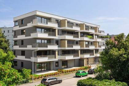 Wohnanlage in der Robert-Stolz-Straße in Linz. Ein modernes, mehrstöckiges Wohngebäude mit hellen Fassaden und zahlreichen Balkonen. Große Fenster sorgen für viel Tageslicht. Vor dem Gebäude stehen Autos auf einer ruhig wirkenden Straße, eingerahmt von gepflegten Grünflächen und Bäumen.