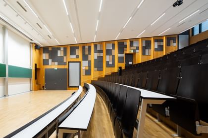 Leerer Hörsaal mit gestuften Sitzreihen und langen Tischen, grüner Tafel, doppelflügeliger Tür und Uhr. Die Wände sind mit Akustikpaneelen in verschiedenen Formen versehen, die Decke mit mehreren Lichtquellen ausgestattet.