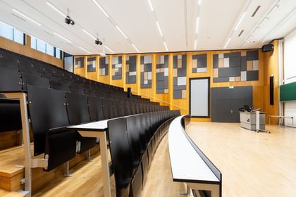 Leerer Hörsaal mit gestuften Sitzreihen und langen Tischen, grüner Tafel, doppelflügeliger Tür und Uhr. Die Wände sind mit Akustikpaneelen in verschiedenen Formen versehen, die Decke mit mehreren Lichtquellen ausgestattet.