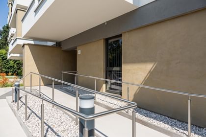 Wohnanlage in der Robert-Stolz-Straße in Linz. Ein modernes, mehrstöckiges Wohngebäude mit hellen Fassaden und zahlreichen Balkonen. Große Fenster sorgen für viel Tageslicht. Vor dem Gebäude stehen Autos auf einer ruhig wirkenden Straße, eingerahmt von gepflegten Grünflächen und Bäumen.