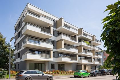 Wohnanlage in der Robert-Stolz-Straße in Linz. Ein modernes, mehrstöckiges Wohngebäude mit hellen Fassaden und zahlreichen Balkonen. Große Fenster sorgen für viel Tageslicht. Vor dem Gebäude stehen Autos auf einer ruhig wirkenden Straße, eingerahmt von gepflegten Grünflächen und Bäumen.