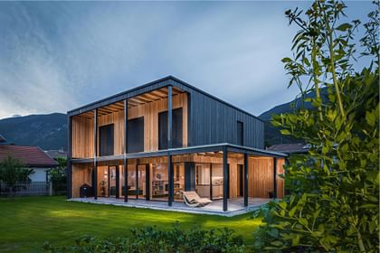 Warm leuchtendes Holzhaus in klarer Kubatur, eingebettet in grüne Geborgenheit