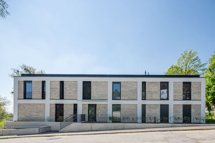 Modernes zweistöckiges Gebäude mit großen Fenstern und Balkonen, mit Bäumen im Hintergrund bei sonnigem Wetter und klarem Himmel.
