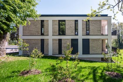 Modernes zweistöckiges Gebäude mit großen Fenstern und Balkonen, gelegen in einer grünen Umgebung mit Bäumen im Hintergrund bei sonnigem Wetter und klarem Himmel.