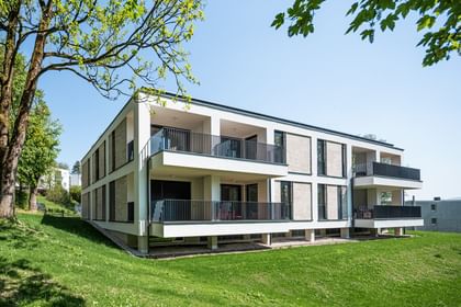 Modernes zweistöckiges Gebäude mit großen Fenstern und Balkonen, gelegen in einer grünen Umgebung mit Bäumen im Hintergrund bei sonnigem Wetter und klarem Himmel.