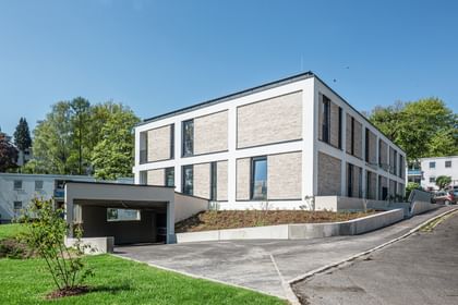 Modernes zweistöckiges Gebäude mit großen Fenstern und Balkonen, gelegen in einer grünen Umgebung mit Bäumen im Hintergrund bei sonnigem Wetter und klarem Himmel.