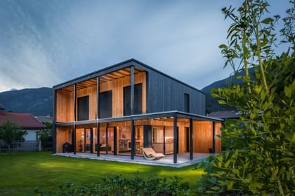 Modernes zweistöckiges Haus mit großen Fenstern und Holzfassade, in der Abenddämmerung beleuchtet. Das Haus ist von grünem Rasen und einem gepflegten Garten umgeben, im Hintergrund sind Berge und weitere Häuser zu sehen.