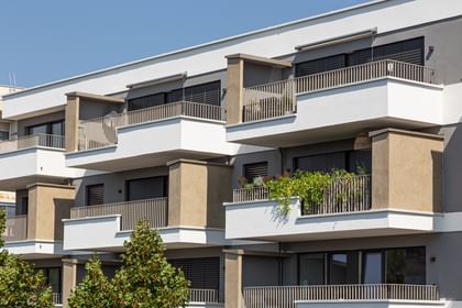 Wohnanlage in der Robert-Stolz-Straße in Linz. Ein modernes, mehrstöckiges Wohngebäude mit hellen Fassaden und zahlreichen Balkonen. Große Fenster sorgen für viel Tageslicht. Vor dem Gebäude stehen Autos auf einer ruhig wirkenden Straße, eingerahmt von gepflegten Grünflächen und Bäumen.
