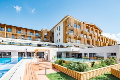 Hotelgebäude mit Holzbalkon und großen Fenstern, das einen Außenpoolbereich umgeben von Grünflächen zeigt. 