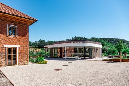 Links ist ein Gebäude mit roten Ziegeln zu sehen, während rechts ein Gebäude mit einer Holzfassade erscheint. Die Umgebung ist grün und der Boden ist mit Kies bedeckt.