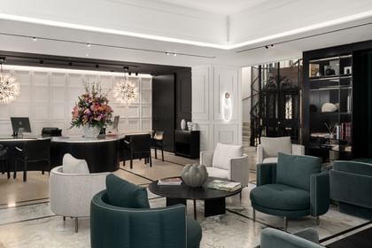 Elegante Lobby im „The Amauris“ Wien mit dunkelgrünen und hellgrauen Lounge-Sesseln um schwarze Couchtische, geschwungenem Empfangstresen mit Blumenarrangement, zwei kugelförmigen Designerleuchten, Marmortreppe neben historischem Aufzug und Regalen sowie weißen Wandpanelen mit indirekter Beleuchtung.
