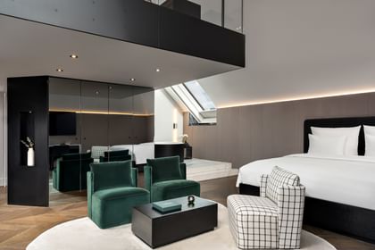 Großzügige Grand-Maisonette-Suite im „The Amauris“ Wien mit Kingsize-Bett, gepolstertem grauen Kopfteil, smaragdgrünen Lounge-Sesseln, kariertem Sessel, schwarzem Couchtisch, Spiegelwand und Galerieebene unter einer Dachschräge mit Oberlicht.