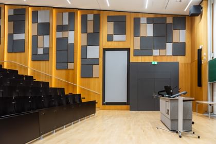 Leerer Hörsaal mit gestuften Sitzreihen, einem Rednerpult mit elektronischer Ausstattung und dekorativen Akustikpaneelen in verschiedenen Formen und Farben an den Holzverkleidungen.