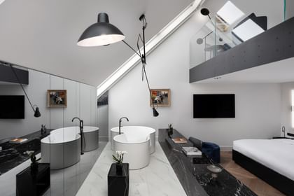 Loftartige Grand-Maisonette-Suite im „The Amauris“ Wien mit zwei freistehenden runden Badewannen auf hellem Marmorboden, langen schwarzen Marmorablagen mit Deko, großem Doppelbett und Galerieebene mit Glasbrüstung unter schräger Dachdecke.