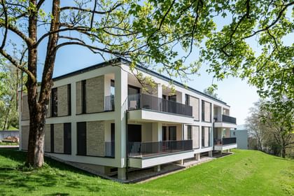 Modernes zweistöckiges Gebäude mit großen Fenstern und Balkonen, gelegen in einer grünen Umgebung mit Bäumen im Hintergrund bei sonnigem Wetter und klarem Himmel.