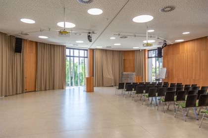 Veranstaltungsraum mit großen Fenstern und Holzwänden. Die Fenster sind teilweise durch Vorhänge bedeckt. Es ist ein Podium zu sehen, vor dem mehrere Reihen schwarzer Stühle stehen.